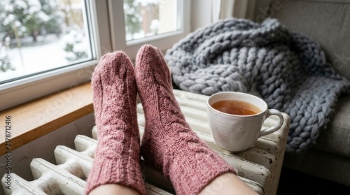 Woman's feet in cozy pink knitted socks resting on a warm radiator with a cup of tea, concept of winter comfort and relaxation
