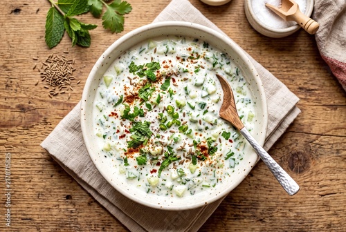 A bowl of creamy Indian raita with cucumber, herbs, and spices on a wooden table.