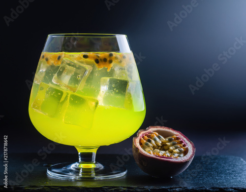 Vibrant yellow refreshing passion fruit cocktail with ice cubes and seeds, beautifully presented beside a halved fruit on a dark, elegant slate surface
