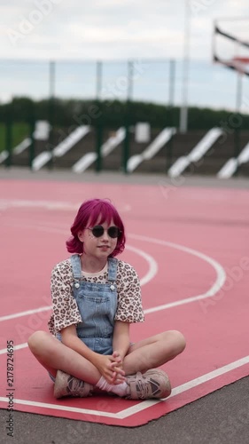 Young girl with short pink hair and straw hat adjusts round sunglasses while sitting crosslegged on red outdoor court marked with white lines, wearing leopard print shirt under denim overalls and