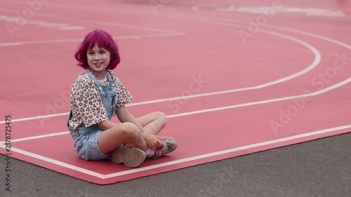 Young child with short pink hair and beige hat sits on red outdoor running track near white curved lane lines, wearing denim overalls with sunglasses clipped to pocket and leopard print shirt