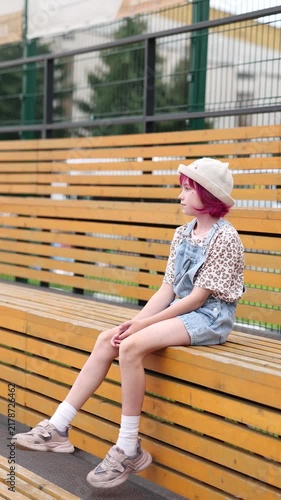 Child with pink hair sits alone on outdoor wooden bleachers wearing light hat, patterned short sleeve shirt, and denim overalls, relaxed neutral expression, casual sneakers and white socks visible
