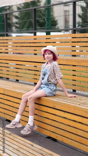 Child with pink hair sits alone on outdoor wooden bleachers wearing light hat, patterned short sleeve shirt, and denim overalls, relaxed neutral expression, casual sneakers and white socks visible