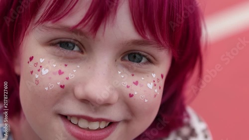 Closeup portrait of smiling child showing upper face and mouth, bright pink dyed hair with bangs framing forehead, tiny heart stickers in white and pink scattered across both cheeks, freckled skin