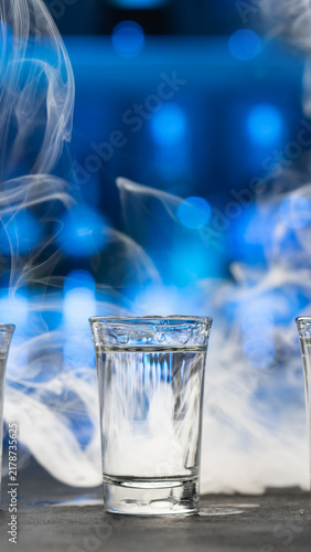 Cold vodka shot in a clear glass surrounded by swirling white smoke and blue bokeh lights. Dramatic vertical beverage composition for alcohol advertising, bars, nightlife and party concepts.