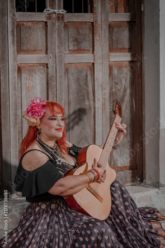 A full-figured Latina Gypsy woman playing traditional Gypsy songs on the guitar; an old yellow house is visible in the background.
