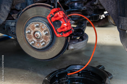 Fotografie Brake repair on car wheel shows red caliper gripping vented disc beside rusted hub and orange hose
