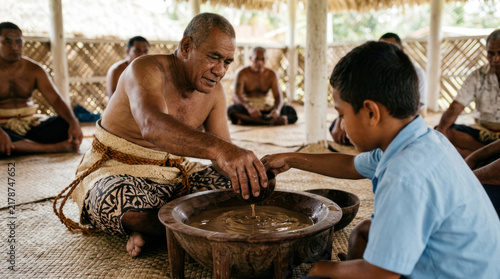 Elder teaches young boy traditional kava ceremony preparation.