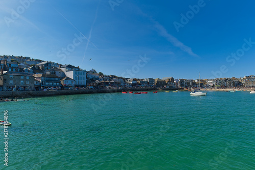 A picturesque Cornish harbor town with turquoise water, boats, and buildings along a hilly coast under a bright blue sky - St Ives, Cornwall, UK