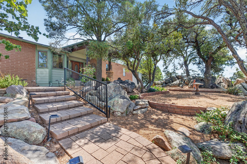 Brick house front yard with stone steps and iron railing, surrounded by trees and rocky garden entry