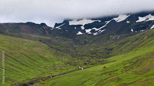 Fotografie A peaceful green valley in northern Iceland with small streams flowing down from snowy cliffs, under low clouds covering the mountain tops
