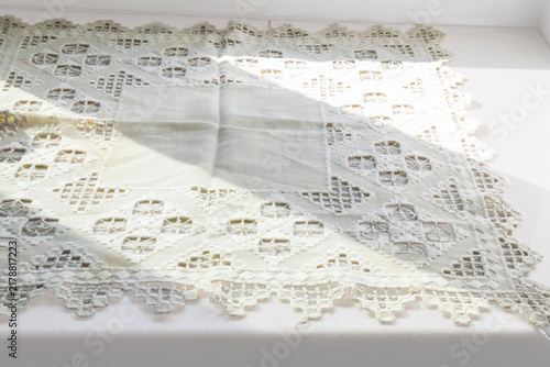 A cream-colored vintage lace tablecloth features intricate openwork embroidery and geometric patterns while draped over a surface under soft natural lighting.