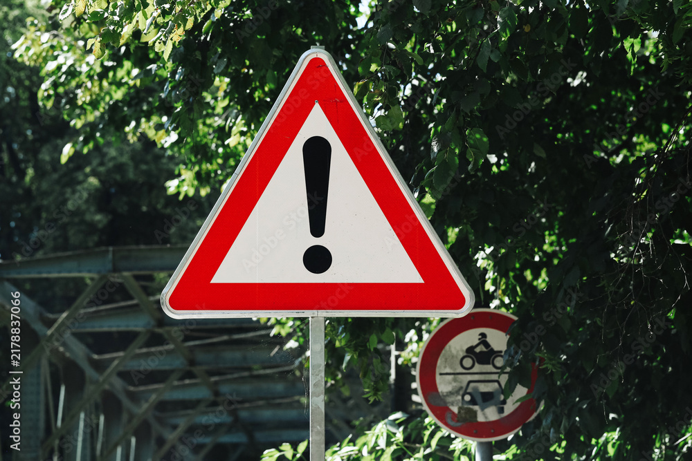 Obraz premium Red triangular warning sign with a black exclamation mark stands beside a road under green trees. General hazard symbol alerts drivers and road users to possible danger and caution background.