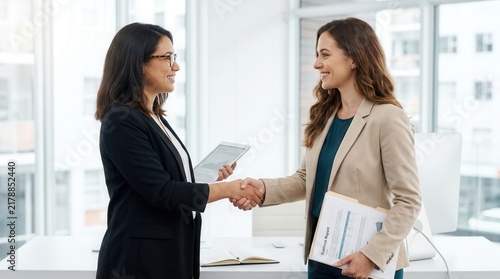 Professional women colleagues from diverse backgrounds shaking hands in a modern coworking office, expressing respect and recognition.