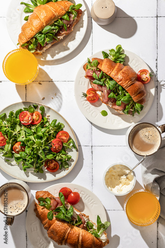 Breakfast table with Croissant sandwiches with prosciutto, cream cheese, corn salad and cherry tomatoes, coffee and orange juice served on white tile table