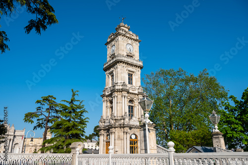 Clock tower with ornate design and white fence in sunny park setting