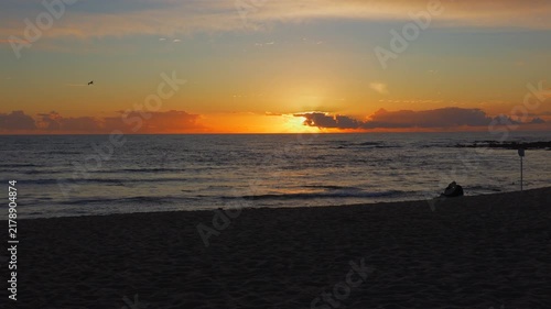Beautiful beach sunset scene in slow motion. A couple sitting on the sand in silhouette as a seagull flies overhead. Vivid read and orange sky.