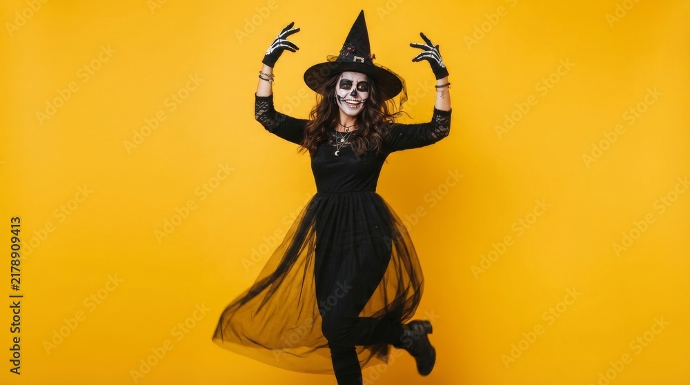 Fototapeta premium Woman in witch hat with sugar skull face paint and skeleton gloves posing dance move against bright yellow studio background