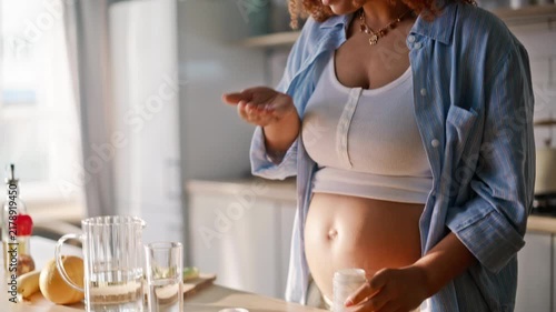 Curly African american pregnant girl taking vitamin pill drinking water kitchen.