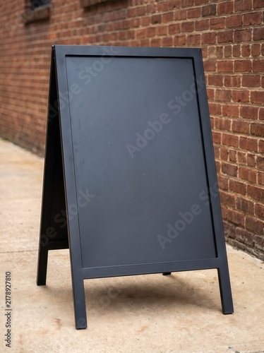 Blank black standing chalkboard sign on pavement, red brick wall background — perfect for cafe, shop, and restaurant branding mockup.