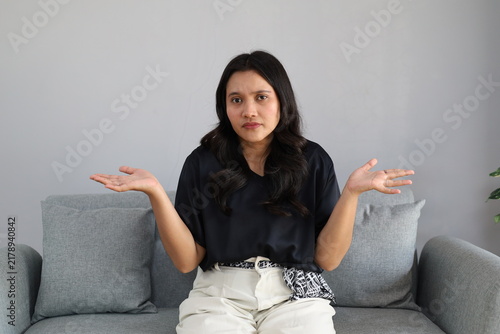 Fotografie A photo of a young woman sitting on the sofa in her living room with a puzzled expression, spreading her hands and shrugging her shoulders