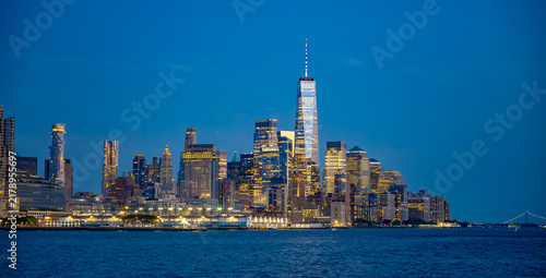 Lower Manhattan skyline and One World Trade Center at blue hour. Panoramic view across New York Harbor shows illuminated towers, piers and the Brooklyn Bridge under a clear sky.