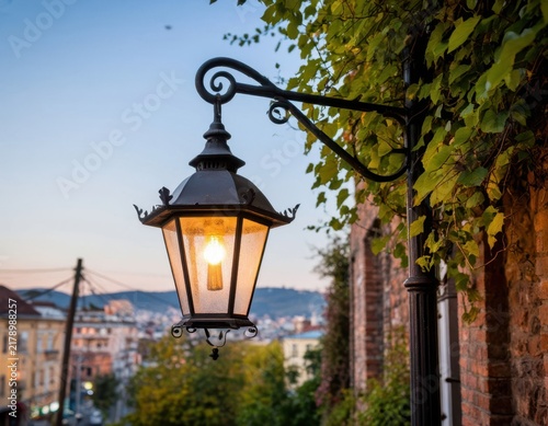 Vintage street lamp illuminated at dusk, surrounded by green vines and brick wall, with cityscape and mountains visible in the background