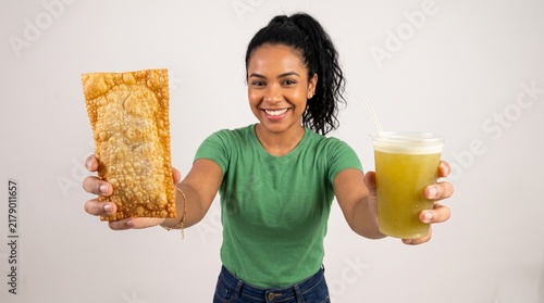 Commercial studio photography cut-out of a cheerful young Brazilian woman extending a crispy golden deep-fried pastel and a cold glass of sugarcane juice towards the camera on an isolated background.