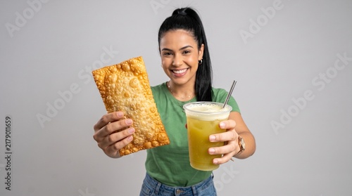 Commercial studio photography cut-out of a cheerful young Brazilian woman extending a crispy golden deep-fried pastel and a cold glass of sugarcane juice towards the camera on an isolated background.
