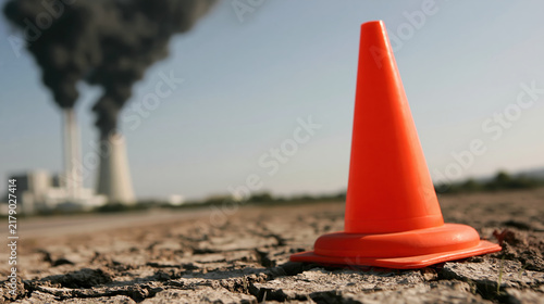Bright orange traffic cone standing alone on cracked dry ground near smoking industrial power plant, creating stark environmental contrast