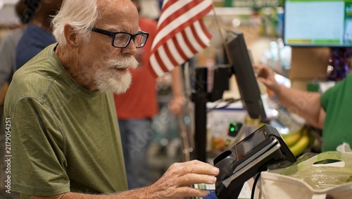 An elderly man is shopping at a grocery store. He is at the checkout counter handling a payment device