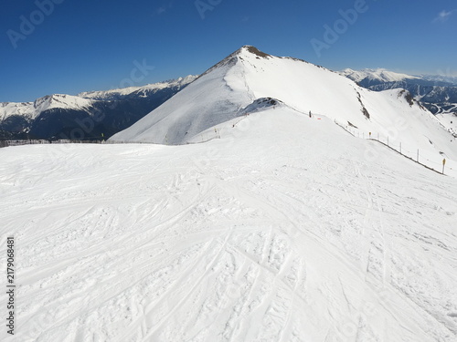 Snow-covered alpine ski resort surrounded by spectacular mountain peaks and valleys under a clear blue sky, with ski slopes, tracks and chairlifts creating a dramatic winter landscape.