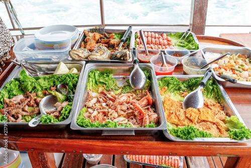 A traditional Thai seafood buffet served on a wooden table on a boat. Includes fried fish, shrimp, calamari, grilled pork, and vegetables with various sauces and sea view background.