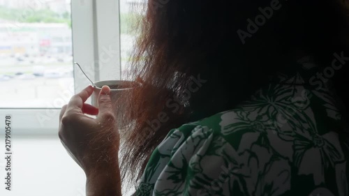 Woman in green patterned clothing holds ceramic cup by large window overlooking urban landscape, captured from behind during quiet morning moment.