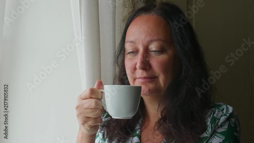 Adult woman sips hot beverage from white ceramic mug indoors near window during morning, peaceful quiet moment at home.