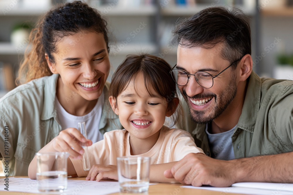Obraz premium Happy family gathers around table, smiling together. Child looks delighted, surrounded by loving parents. Glasses and casual clothes show relaxed, joyful mood. Perfect for family bonding, education