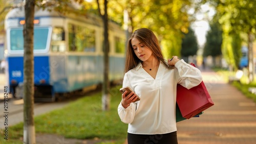 Bild auf Leinwand Young woman with shopping bags checks her phone while walking near a tram