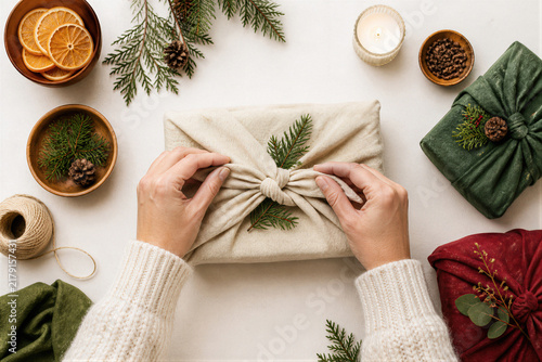 Woman hands wrapping Christmas gift in fabric using furoshiki technique. Eco friendly holiday present with evergreen branch decoration.
