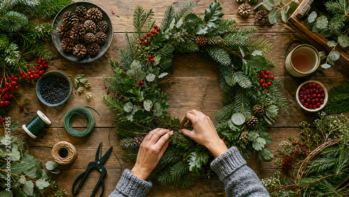 Woman hands making christmas wreath from evergreen branches on rustic wooden table. Diy festive decoration for winter holiday season. Craft workshop with pine cone and holly berry.