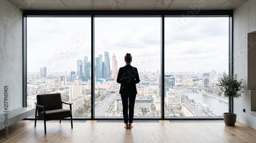 Silhouette of confident businesswoman standing alone in front of large floor to ceiling window looking out at panoramic city skyline view with skyscrapers and river