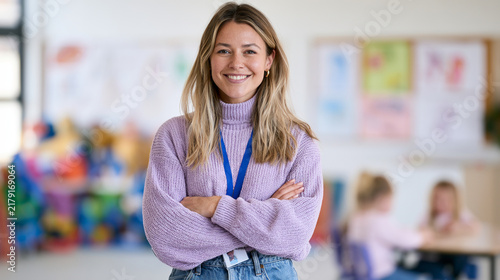 Smiling blonde female teacher stands with crossed arms in a colorful classroom. Early education, teacher portrait, school staff, learning environment.