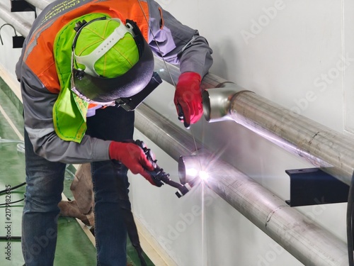 A medium shot of an industrial worker bending over to perform precise TIG welding on a wall-mounted stainless steel pipe line, producing a bright arc glare while wearing a flip-up face shield