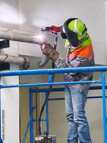 A low-angle shot of an industrial welder working on scaffolding, using TIG welding on a stainless steel pipe joint, creating a bright welding arc light flare while wearing a flip-up welding mask