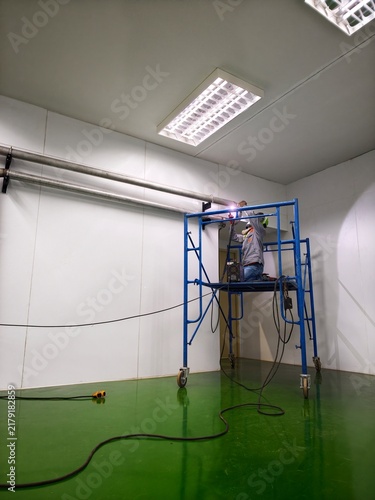 A wide-angle, low-angle shot of a technician standing on mobile scaffolding to perform pipe welding near the ceiling in a spacious cleanroom facility with a green epoxy floor 