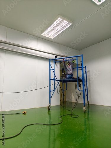 A wide-angle, low-angle shot of a technician standing on mobile scaffolding to perform pipe welding near the ceiling in a spacious cleanroom facility with a green epoxy floor 