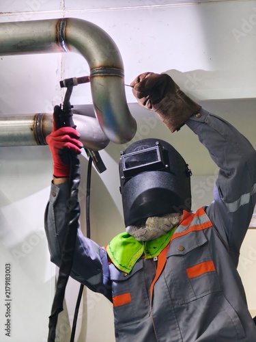 A low-angle shot of a professional welder wearing a welding helmet and protective gloves performing TIG welding on an overhead stainless steel pipe joint in an industrial facility.