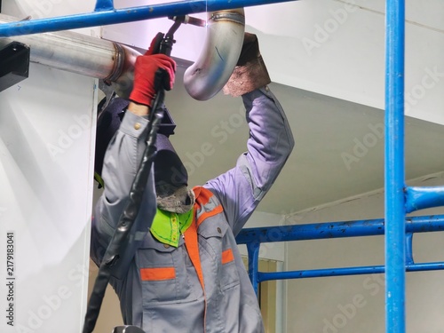 A low-angle shot of a professional welder wearing a welding helmet and protective gloves performing TIG welding on an overhead stainless steel pipe joint in an industrial facility.
