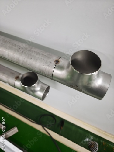 A close-up shot of a welded stainless steel tee fitting connected to stainless steel piping, showing heat discoloration along the circumferential weld seams and printed specification markings on the f