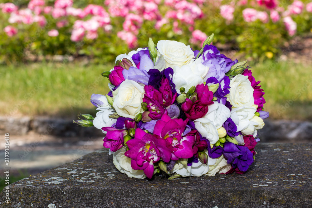 Obraz premium Colorful wedding bouquet resting on stone with roses in background