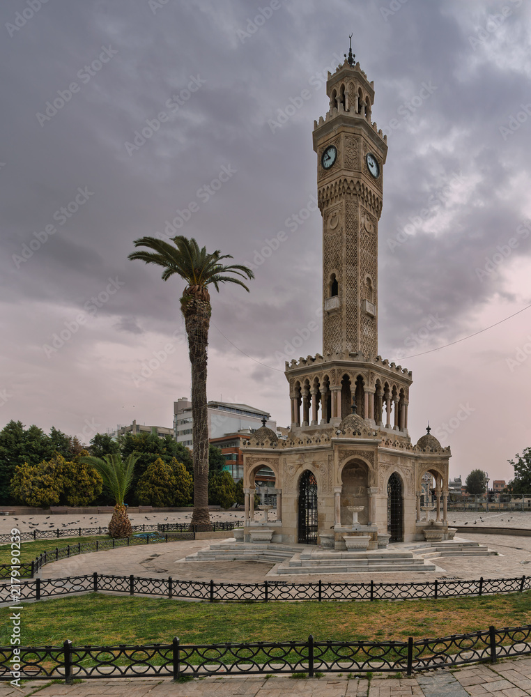 Fototapeta premium Historic Izmir Clock Tower at Konak Square in Izmir, Turkey, an iconic Ottoman landmark featuring intricate stone carvings and a prominent palm tree under a dramatic cloudy sky